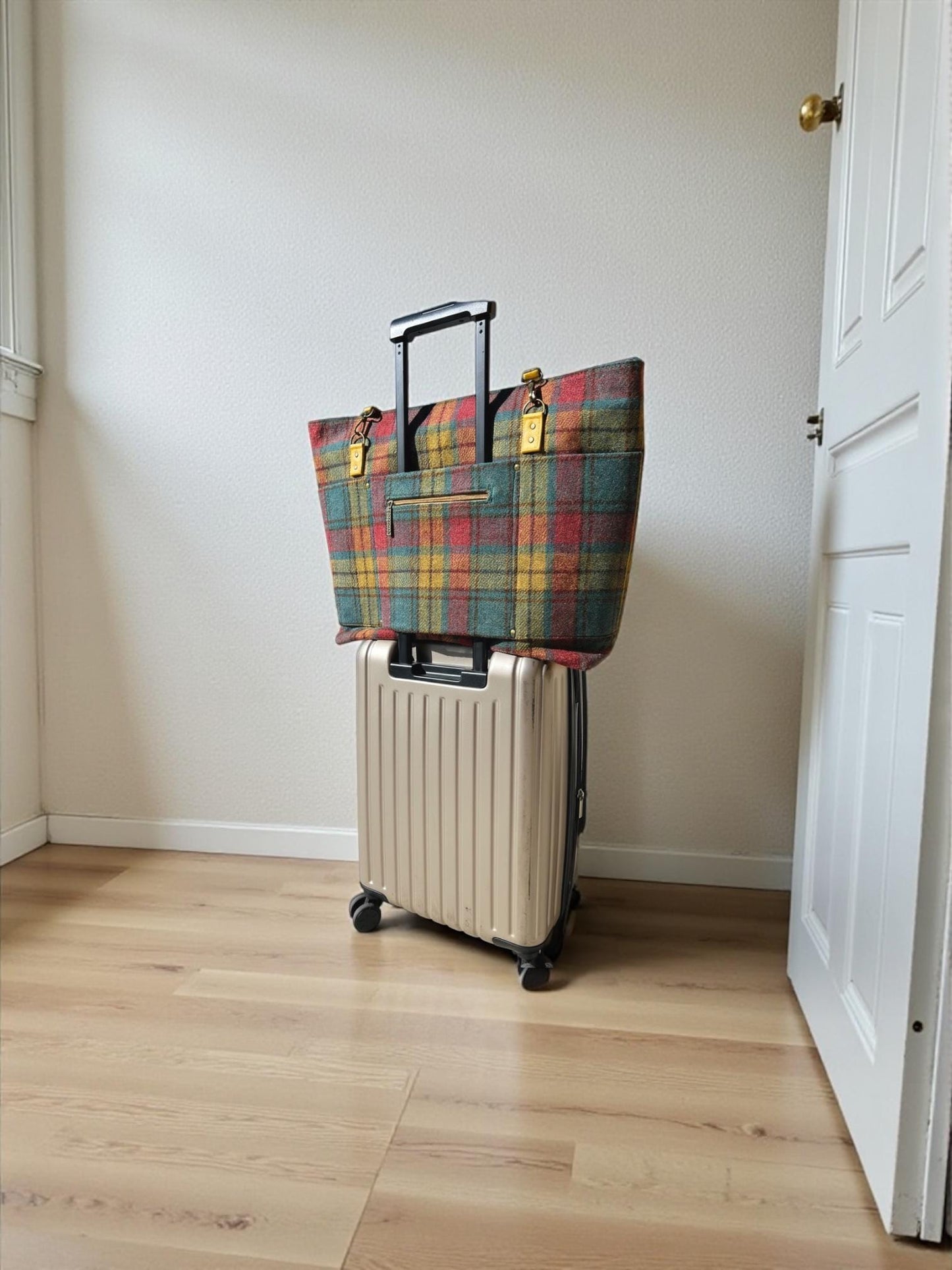 An extra large turquoise, fuchsia, yellow and dark brown plaid Harris tweed tote bag sitting atop a carry-on roller bag with the suitcase handle through the trolley sleeve. The trolley sleeve has a zippered pocket with a Celtic knot antique bronze pull. The sides of the tote extend past the carry-on bag by approximately 3 inches on each side. The goldenrod colored strap connectors are attached with antique bronze rivets and fitted with antique bronze d rings and swivel hooks on the straps.