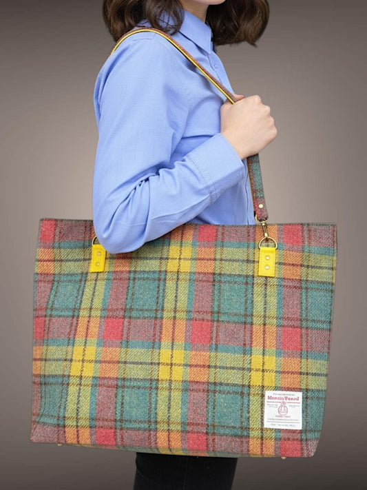 A woman in a collared  blue business shirt holds a giant Harris Tweed bag on her shoulder.  The bag is turquoise, fuchsia, and goldenrod yellow plaid with dark brown checks. The authentic Harris Tweed Authority Label is in the lower right corner of the front of the bag. The two sided strap has the golden rod faux leather peeking out from the bottom of the Harris Tweed  top side. Golden rod strap connectors are attached to the bag with stitching and reinforced with two rivets on each connector.