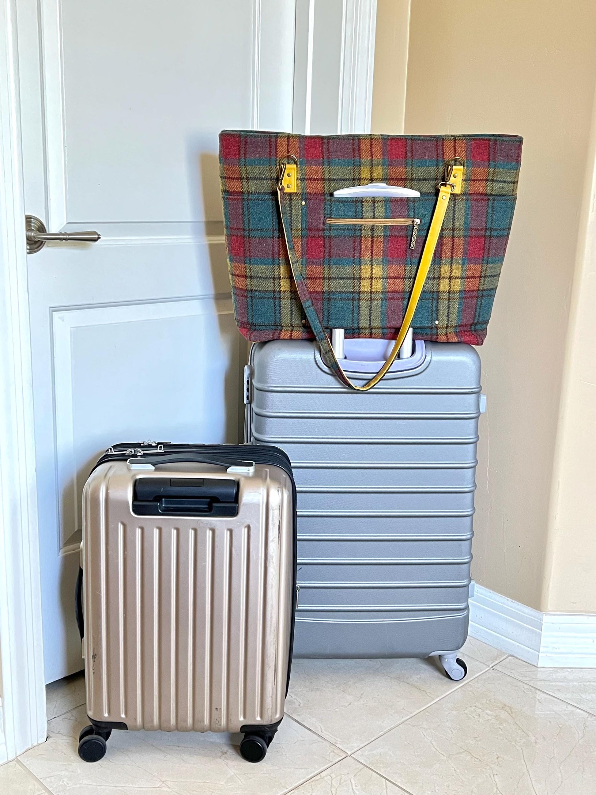 The extra large Harris tweed tote is set a top a large check-in roller bag with a small carry-on roller bag next to it. The sides of the tote only extend past the top of the large check-in roller bag by an inch on each side. The handle of the large check-in bag fits through the trolley sleeve. The strap of the tote hangs down so that the two-sided strap can be seen with Harris Tweed on the top and a golden rod faux leather on the bottom.